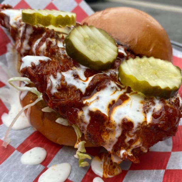 A B-UNOS spicy fried chicken sandwich with pickles, sauce, and lettuce on a red and white checkered paper.