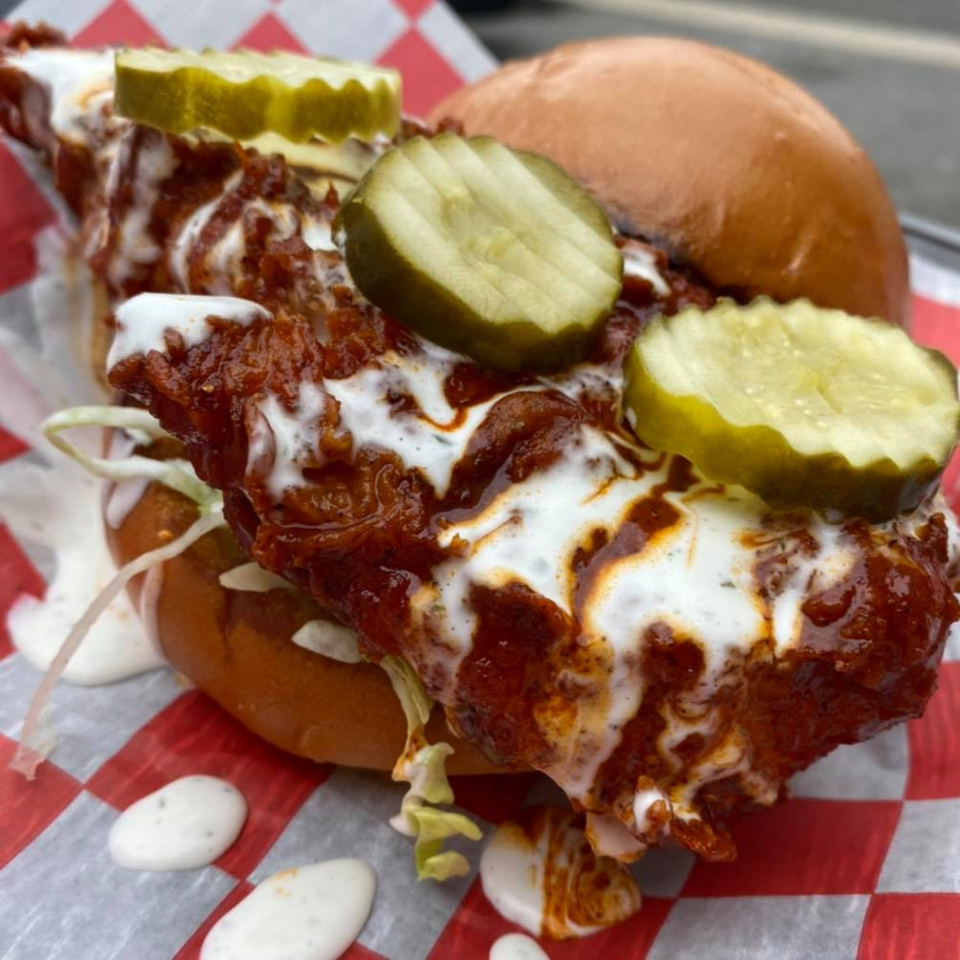 A B-UNOS spicy fried chicken sandwich with pickles, sauce, and lettuce on a red and white checkered paper.