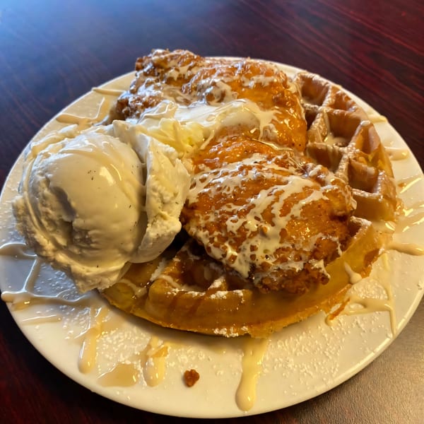 Chicken and waffles with vanilla ice cream, syrup, and powdered sugar on a white plate.