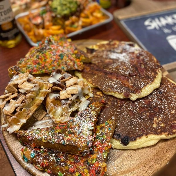 A wooden board with pancakes, waffles, and French toast covered in cereal and powdered sugar.