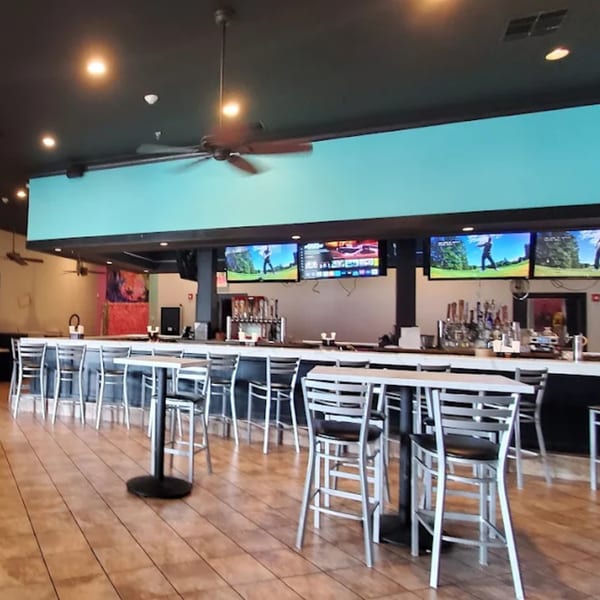 Interior of B-UNOS restaurant with a long bar, high-top tables, chairs, and TVs showing golf.