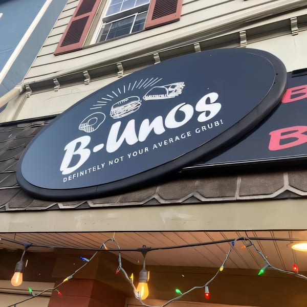 B-UNOS restaurant sign, a dark oval with white text and food illustrations, above string lights.