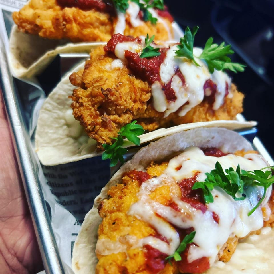 Three B-UNOS fried chicken tacos with melted cheese, red sauce, and parsley on a metal tray.