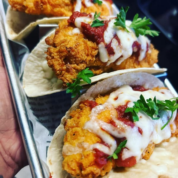 Three B-UNOS fried chicken tacos with melted cheese, red sauce, and parsley on a metal tray.