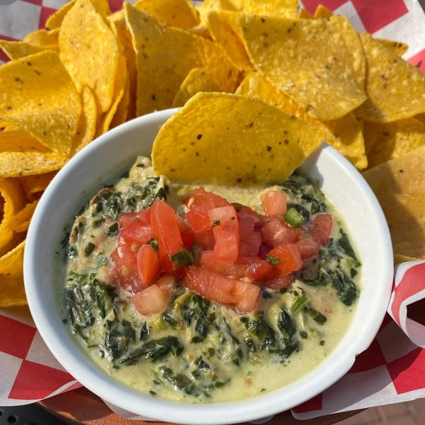 Spinach dip topped with diced tomatoes, served with tortilla chips on a red and white checkered liner.