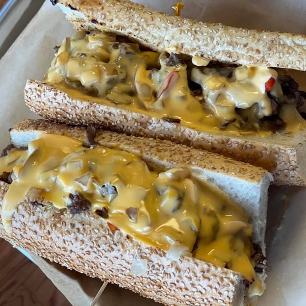 Two B-UNOS cheesesteak sandwiches, overflowing with melted cheese, steak, and onions on sesame seed rolls.