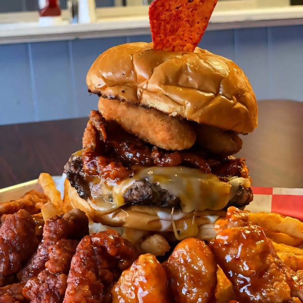 A large B-UNOS burger with melted cheese, onion rings, and a chip, surrounded by fries and wings.